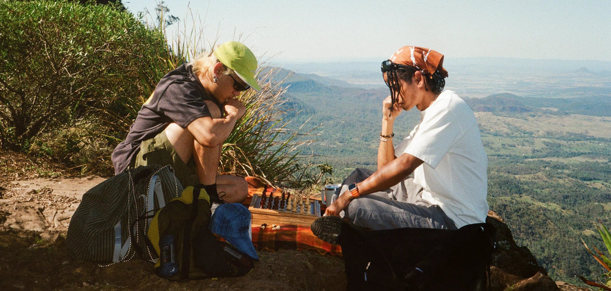 Two friends playing chess on a sunny mountain lookout, wearing Gramicci
